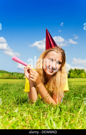 Wunderschönes kleines Mädchen mit Krachmacher Horn und Geburtstag party Kappe Verlegung außerhalb mit glücklichen Ausdruck an sonnigen Sommertag Stockfoto