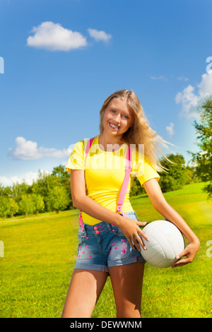 Glücklich und lächelnd 13 Jahre altes Mädchen mit langen blonden Haaren stehen auf der Wiese mit dem Ball im Park an sonnigen Sommertag Stockfoto