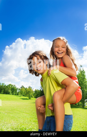 Zwei schulpflichtige Kinder Jungen tragen Mädchen auf dem Rücken, beide glücklich, Lächeln, auf sonnigen Tag Stockfoto