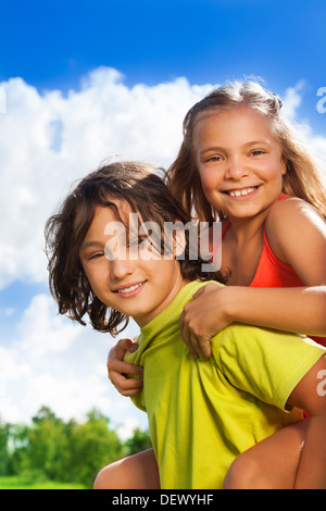 Enge Portrait zwei schulpflichtige Kinder Jungen tragen Mädchen auf dem Rücken, beide glücklich, Lächeln, auf sonnigen Tag Stockfoto