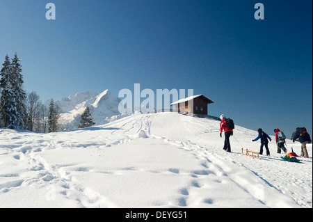 Rodler und ein Holzhaus auf einem schneebedeckten Hügel Mt Alpspitze und Mt Zugspitze am Rücken, Eckbauer, Garmisch-Partenkirchen Stockfoto