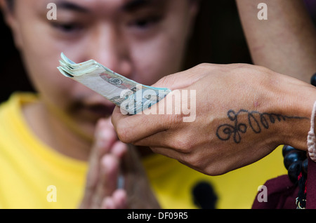 Charme-Verkauf, Mann, der die traditionelle Geste von Gruß und zeigen Respekt, Wai, tätowiert Hand Holding Banknoten, Tempel Stockfoto