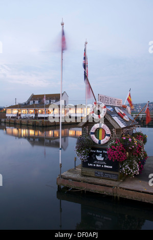 Blumen und Fahnen schmücken die Ticket-Hütte in West Bay River Boot mieten. Über das Wasser ist das Riverside Restaurant. Dorset, England, Vereinigtes Königreich. Stockfoto