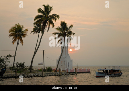 Ein Boot, vorbei an einer Kirche bei Sonnenuntergang, am Ufer des Vembanad See auf den Backwaters von Kerala, Kumarakom, Indien, Asien Stockfoto