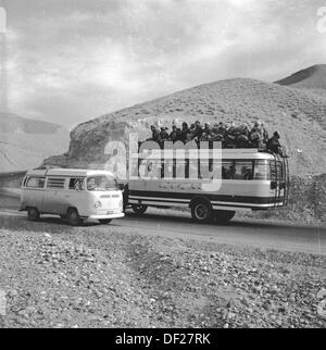 Historisches Bild aus den 1950er Jahren, zeigt einen voll beladenen afghanischen Bus - mit Fahrgästen drinnen und auf dem Dach - und einen VW-Wohnmobil, der auf einer Schotterstraße in einer bergigen Gegend Afghanistans fährt. Stockfoto