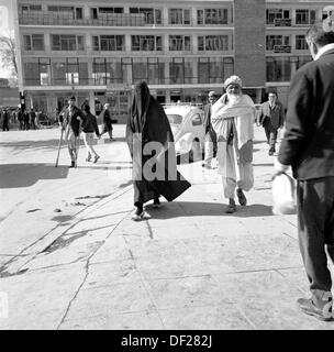 1950er Jahre, historisches Bild von J Allan Cash eines älteren afghanischen Mannes, der neben einer vollständig verschleierten Dame in einer Straße in Kabul, Afghanistan, geht. Diese Ära war eine relativ friedliche Zeit für das Land, in der eine Reihe neuer, moderner Gebäude errichtet wurden, wie auf dem Bild zu sehen ist und Anstrengungen unternommen wurden, um das Land zu modernisieren und eine offenere, wohlhabendere Gesellschaft zu schaffen. Dies endete in den 1970er Jahren mit Invasionen, Staatsstreichs und Bürgerkriegen. Stockfoto