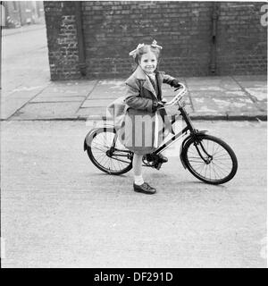 Geschichtsbild, der ein süßes kleines Mädchen auf einem großen Fahrrad in eine leere Stadtstraße zur Schule, England der 1950er Jahre. Stockfoto