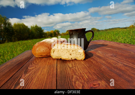 Selbstgebackenes Brot und Becher Milch auf einem Holztisch. Hintergrund blauer Himmel. Outdoor-Farm-Stil Stockfoto