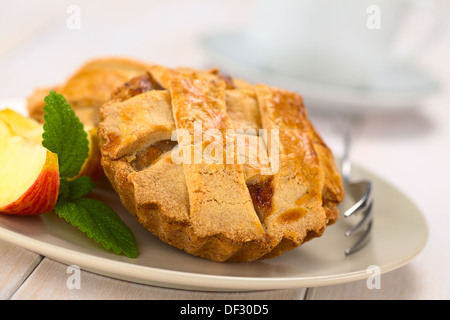 Kleine Runde Apfelkuchen mit Gitter-Kruste mit Kuchengabel, Minze und Apfel Scheiben auf einem Teller, Tasse in den Rücken Stockfoto