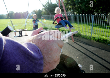 Mann Rauchen in einen Kinderspielplatz mit einem kleinen Mädchen auf einer Schaukel mit den Händen auf einen buggy Stockfoto