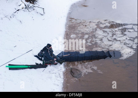 Winter schnee Szene von Ufer von teilweise zugefrorenen Fluss wharfe - 1 Mann sitzt auf Kiste, Angeln mit Rute, Line & große keepnet - Skipton, England, UK. Stockfoto