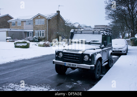 Fahrzeuge (einschließlich eines Land Rover Defender) an der Seite der Straße in einem Wohngebiet geparkt, an einem verschneiten Tag. Stockfoto