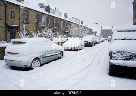 Schneebedeckte Autos entweder Seite des engen Wohn- Urban Street auf verschneiten Wintertag geparkt, Reifenspuren, die zur Mitte der Straße - Baildon, Yorkshire, Großbritannien. Stockfoto