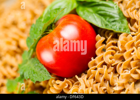 Geneigt Closeup horizontale Foto einzelne reife Tomate, umgeben von geschweiften Teigwaren Nudeln und großes Blatt Basilikum Stockfoto