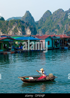 Eine Vietnamesin in einem konischen Hut Zeilen ein Boot vorbei an einem schwimmenden Dorf von Cat Ba Insel im Lan Ha Bucht, Halong Bucht, Vietnam. Stockfoto