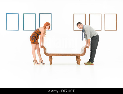 zwei kaukasische Menschen, die versuchen, eine Holzbank in einem weißen Raum mit leere Frames angezeigt an Wänden zu erheben Stockfoto