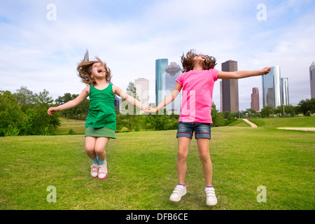 Zwei Schwester Mädchen Freunde springen glücklich halten hand in urbanen moderne Skyline Park Rasen Stockfoto