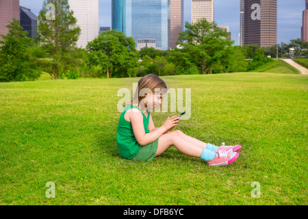 Blonde junge Mädchen spielen mit Smartphone auf Park Rasen in Stadt Skyline Hintergrund sitzen Stockfoto