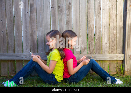 Zwillingsschwester Mädchen spielen mit tablet-pc auf Hinterhof Rasen Zaun gelehnt auf dem Rücken sitzen Stockfoto