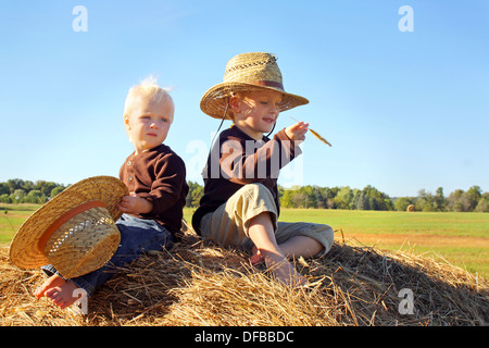 Zwei kleine Kinder, einen kleinen Jungen und seinem kleinen Bruder, sitzen auf einem Heuballen tragen Strohhüte an einem sonnigen Herbsttag. Stockfoto