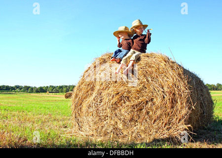 Zwei glückliche Kinder, einen jungen und seinen kleinen Bruder, sitzen auf einem Heuballen in einem Feld auf einem Bauernhof, Stroh-Cowboy-Hüte tragen. Stockfoto