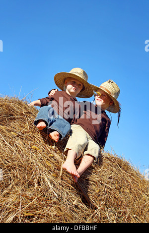 Zwei glückliche Kinder, einen jungen und seinen kleinen Bruder, sitzen auf einem Heuballen in einem Feld auf einem Bauernhof, tragen Cowboy Strohhüte Stockfoto