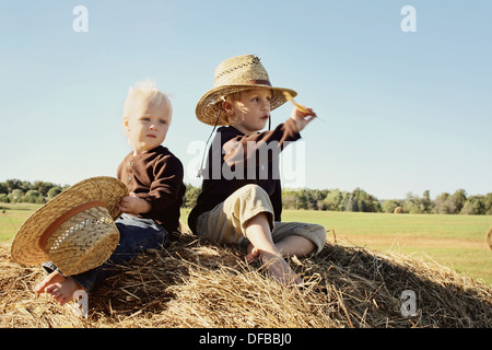 Zwei glückliche kleine Jungs, 4 Jahre alt und Baby Bruder sind sitzt oben auf einem großen Heuballen, tragen Strohhüte auf Herbsttag Stockfoto