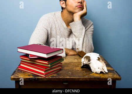 Junger Mann sitzt an einem Schreibtisch mit einem Stapel von Büchern und ein Tier Schädel Stockfoto