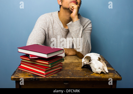 Junger Mann sitzt an einem Schreibtisch mit einem Stapel von Büchern und ein Tier Schädel Stockfoto