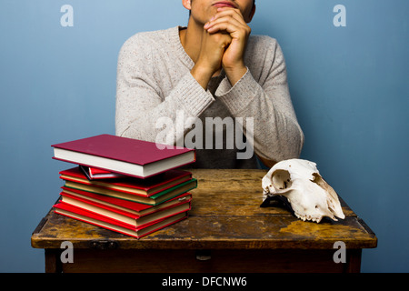 Junger Mann sitzt an einem Schreibtisch mit einem Stapel von Büchern und ein Tier Schädel Stockfoto