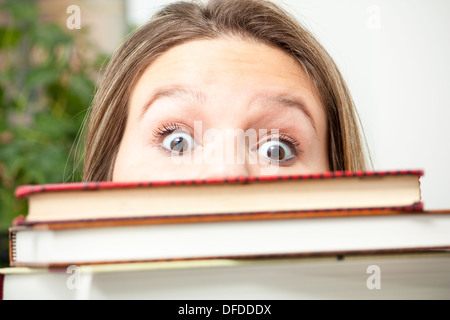 Eine junge Studentin guckt sich über einen großen Stapel Bücher in Panik über die Hausaufgaben, die sie zu tun hat. Stockfoto