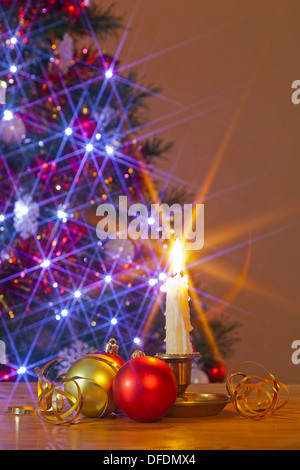Christbaumschmuck Christbaumkugel und eine Kerze auf einem Tisch vor einem geschmückten Baum mit Lichterketten. Stockfoto