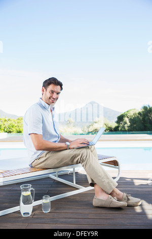 Mann mit Laptop auf Liegestuhl am Pool Stockfoto