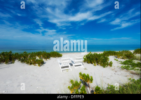 Zwei weiße hölzerne Liegestühle im Sand mit Blick auf den Ozean Meer Ufer unter sonnigen warmen blauen Himmel blau-grünen Ozean Stockfoto