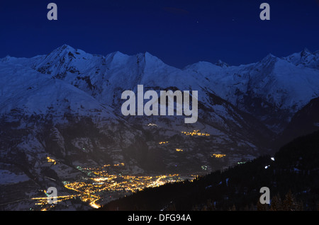 Die französischen Alpen des Tals Tarantaise sind bei Mondschein beleuchtet, wie die Straßenlaternen von Bourg Saint Maurice unten leuchten. Stockfoto