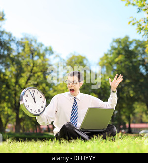 Junge wütend Unternehmer mit Laptop auf dem grünen Rasen sitzen und Blick auf die Uhr in einem park Stockfoto