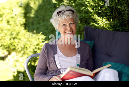Ältere Frau saß auf einem Stuhl im Garten ein Buch hält und mit Blick auf die Kamera zu Lächeln Stockfoto