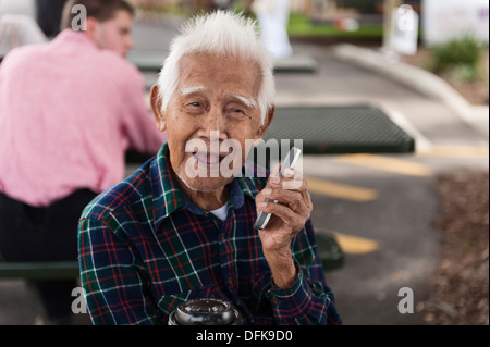 Ein Senioren-Mann ohne Zähne halten ein smart Phone nah an sein Gesicht sitzend auf einer Bank am Wooton Park, Tavares FL Stockfoto