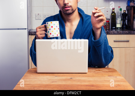 Junger Mann im Bademantel mit Laptop und Rauchen Stockfoto