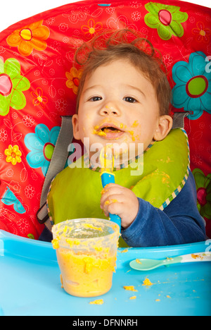 Chaotisch Baby Püree mit Löffel essen, von sich selbst und sitzen Stuhl Stockfoto