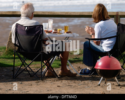 Älteres Paar mit einem BBQ auf einer Klippe mit Blick auf den Strand, Widemouth Bay, Cornwall, UK Stockfoto