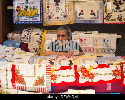 Portugal Algarve Loule Markt nachdenklich gelangweilt suchen faltig alt grau grauhaarige Dame Frau Kopftuch Souvenir Küche Tee Handtuch Display Stand Stockfoto