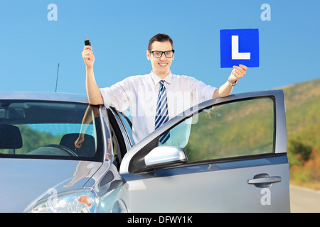 Glücklich Jüngling posiert in der Nähe von seinem Auto hält einen L Zeichen und Schlüssel, nachdem er seinen Führerschein auf einer Straße Stockfoto