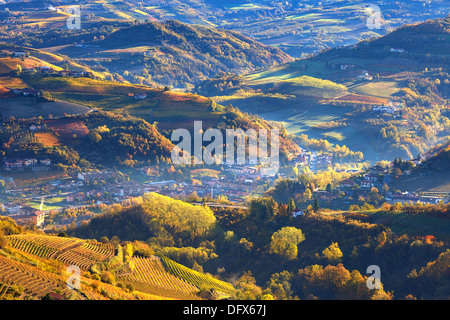 Nebligen Morgen über Kleinstadt unter herbstlichen Hügeln und Weinbergen des Piemont, Norditalien (Ansicht von oben). Stockfoto