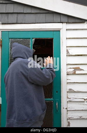 Eine verdächtige Person, Mann, trägt eine Kapuzenjacke spähte durch eine zerbrochene Fensterscheibe in einer Tür ein Gartenhaus. Stockfoto