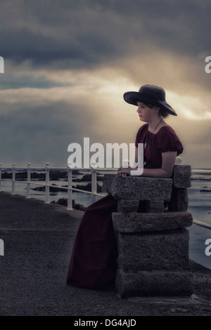 ein trauriges Mädchen mit einer schwarzen Sonnenhut sitzt auf einer Steinbank am Meer Stockfoto