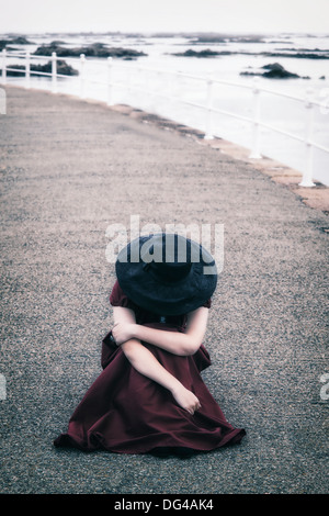 eine Frau in einem roten Kleid mit einer schwarzen Sonnenhut sitzt auf einem pier Stockfoto