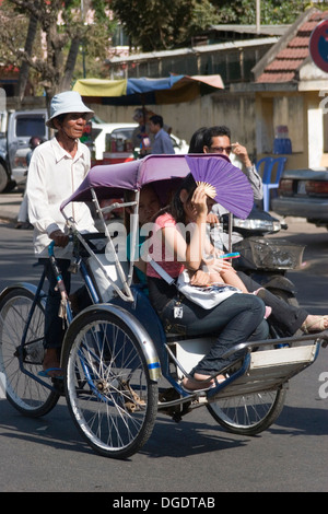 Ein Mann ist ein Cyclo mit einem Passagier hausieren, die sich mit einem Ventilator auf einer Stadtstraße in Phnom Penh, Kambodscha Schattierung ist. Stockfoto