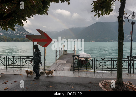 Weibliche junge Erwachsene zu Fuß ihren beiden Hunden vorbei ein Regenbogen über Lago di Lugano, Tessin, Schweiz Stockfoto