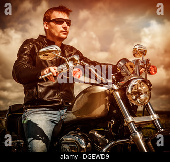 Biker-Mann trägt eine Lederjacke und eine Sonnenbrille sitzt auf seinem Motorrad, Blick auf den Sonnenuntergang. Stockfoto